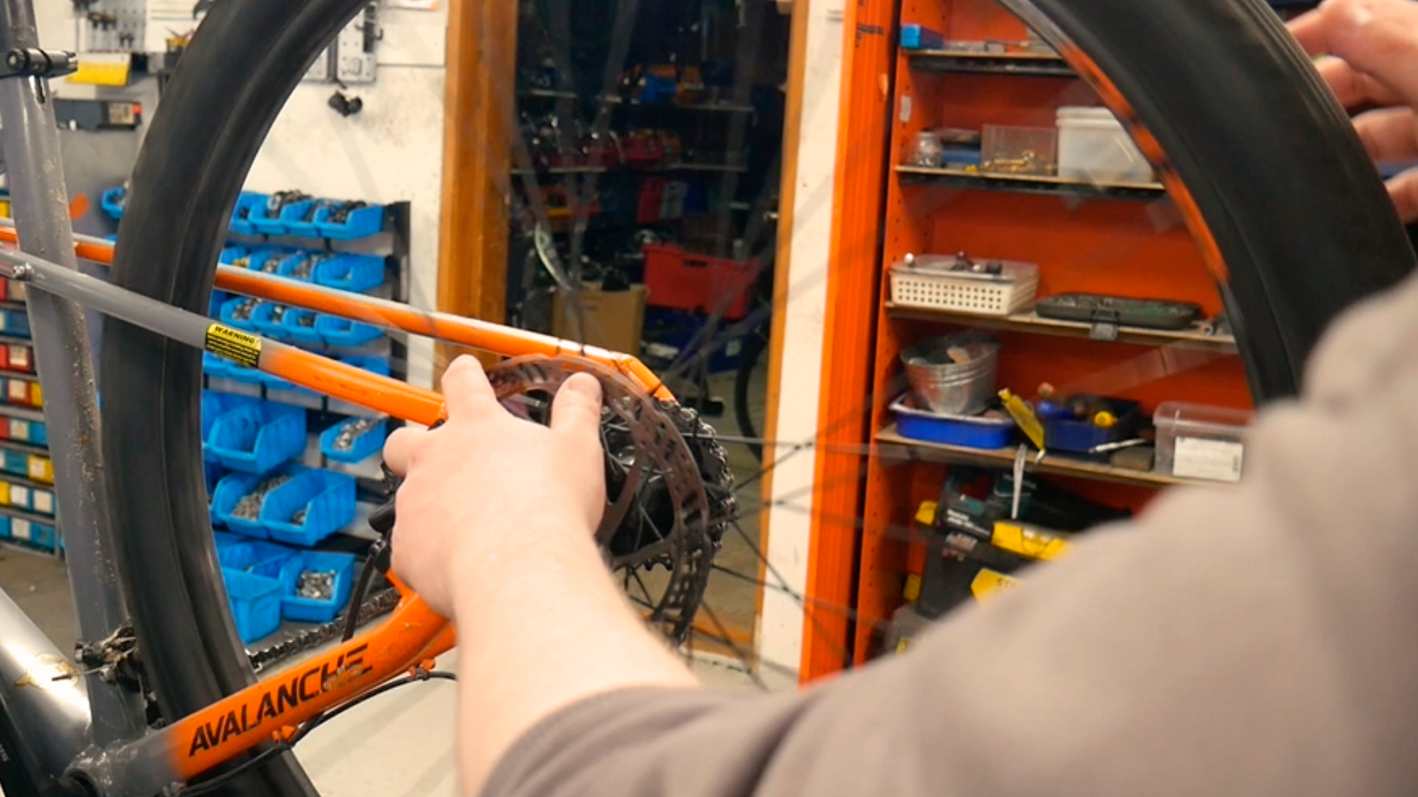 Pyöräkorjaamolla korjataan oranssinharmaata Avalanche-merkkistä polkupyörää. Renkaiden pinnojen välistä näkyy korjaamon tilat. Seinähyllyt ovat täynnä työkaluja ja varaosia.