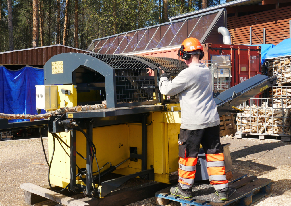 Henkilö pilkkoo polttopuita leikkuukoneella kestävän kehityksen keskuksen pihalla.