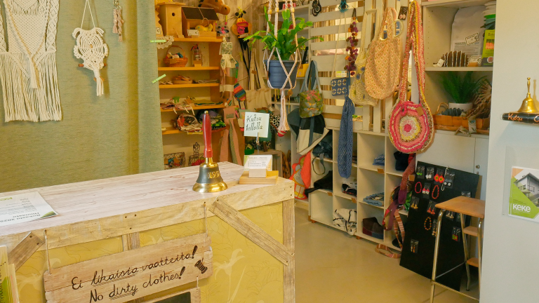 Kestävän kehityksen keskuksen myymälän myyntipiste. Etualalla on tiski, jossa on soittokello, käyntikorttiteline, sekä lapulla kehotus "Kutsu kellolla". Tiskin eteen on kiinnitetty kyltti, jossa lukee "Ei likaisia vaatteita! No dirty clothes!". Tiskin takaa avautuu näkymä myymälähyllyihin, joissa näkyy lukuisia myytäviä tuotteita.