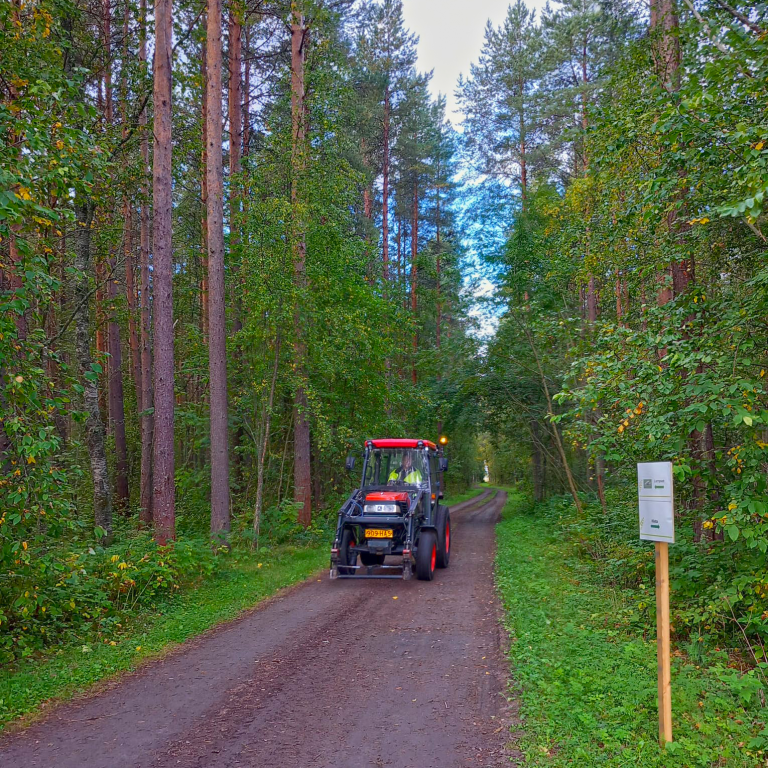 Hietasaaren metsäpolulla työvaatteisiin pukeutunut henkilö kulkee traktorilla kohti Hietan toimipaikkaa. Tienviitta osoittaa, että Hietan lisäksi polkua toiseen päähän kulkemalla voi päästä katsomaan lampaita.
