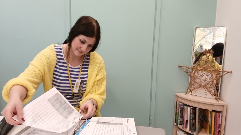 Työllisyyspalvelupäällikkö Rosalie Leskelä laittamassa lomaketta mappikansioon. Toimiston nurkassa on pieni kirjahylly, jonka päälle on asetettu punottu tähtikoriste ja peili.