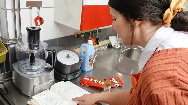 Työllisyyspalvelupäällikkö Rosalie leskelä lukemassa Omakupin keittiössä reseptikirjaa. Pöydällä näkyy soseutin, muita keittiövälineitä, sekä avattu paketti taloussuklaata ja nonparelleja leivontaa varten.