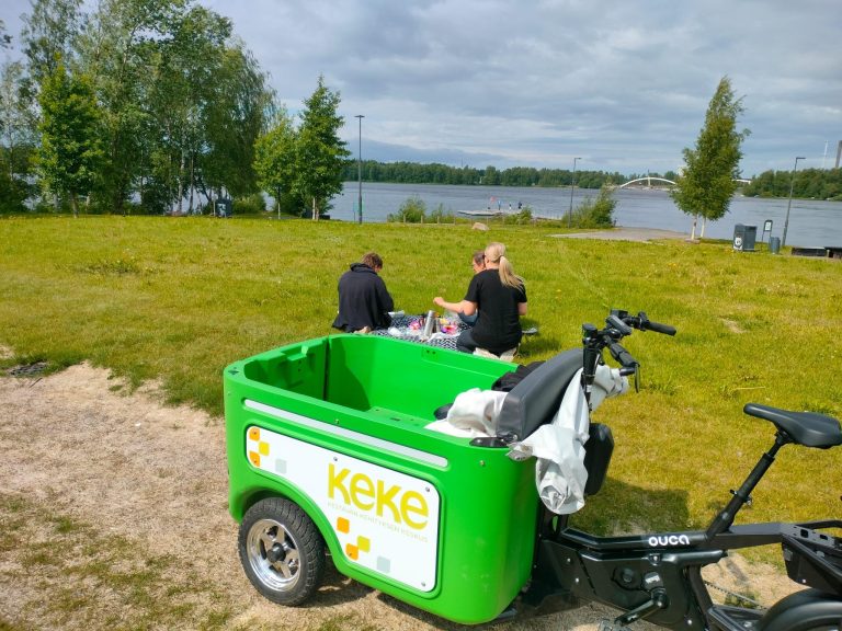 Ouca Bikesin keke-brändätty sähköpyörä on pysäköity nurmikon viereen. Pyörän tilava tavaratila on auki ja tyhjillään. Nurmikolla näkyy Kestävä elämä -hankkeelaisia piknikillä, ihastelemassa taustalla näkyvää vesimaisemaa.