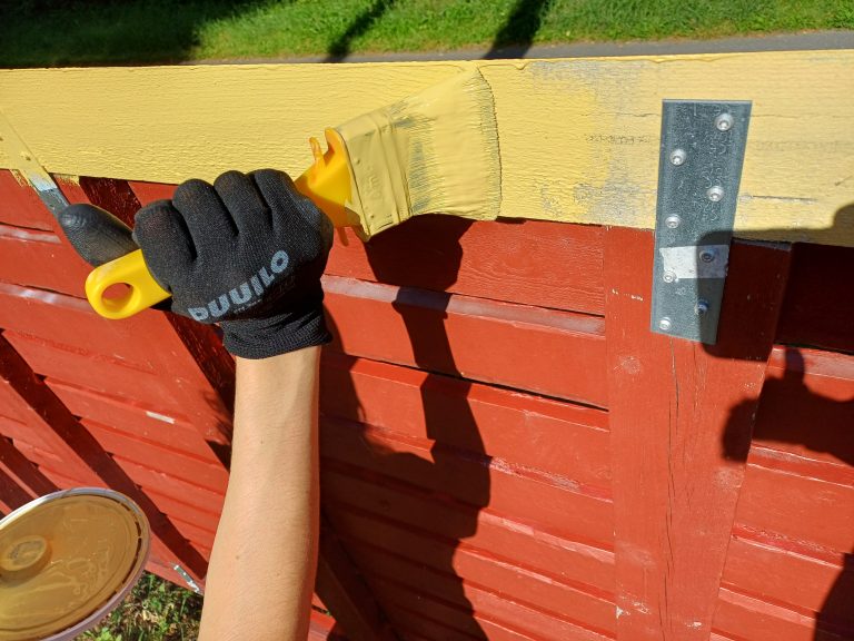 Hansikkaat kädessään oleva henkilö maalaamassa aitaa.