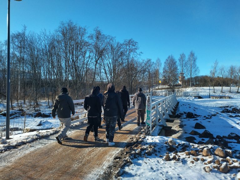 Henkilöitä talvivaatteissa ylittämässä puron yli kulkevaa siltaa talvisessa Oulun puistossa.