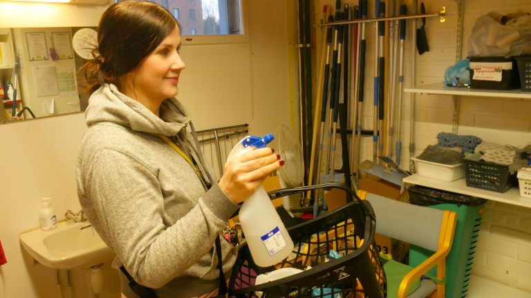 Työllisyyspalvelupäällikko Rosalie Leskelä on puhtaanapidon palvelun tiloissa. Huoneen seinillä on moppeja, lastoja ja hylly täynnä siivousrättejä. Rosalie pitelee oikeassa kädessään suihkepulloa täynnä pesuainetta, ja vasemmassa kädessään koria täynnä erinäisiä siivoustarvikkeita.