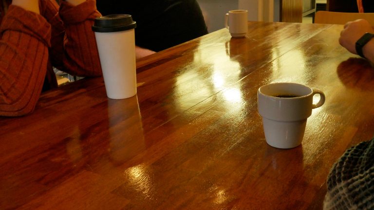 Hämärästi valaistu lounasruokala Omakupin ruokapöytä. Pöydän ääressä istuu mikrotuen henkilöstöä ja Rosalie. Pöydällä on kahvikuppeja.