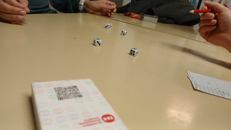 Pöydän ääreen on kerääntynyt joukko ihmisiä. Pöydällä on Yatzy-noppapelin pakkaus, viisi noppaa ja pisteidenlaskuvihkonen. Yhdellä henkilöllä on kädessään kynä pisteiden merkkausta varten. Pöydän toiseen päähän on asetettu Uno-korttipeli.