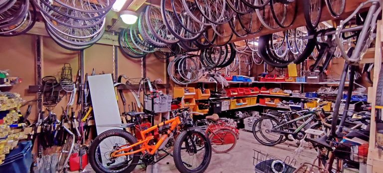 Pyöräkorjaamon tarvikevarasto. Seinät ovat täynnä hyllyjä ja telineitä erinäisille varaosille. Katosta roikkuu renkaita ja vanteita. Lattialla on kunnostettavana olevia polkupyöriä.