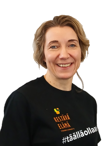 Hankekoordinaattori Päivi Parkkila hymyilee. Hänellä on päällään tumma paita, jossa on Kestävä elämä -hankkeen logo ja teksti "hashtag täällä ollaan".