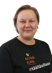 Hankepäällikkö Ulla Heikkilä hymyilee. Hänellä on päällään tumma paita, jossa on Kestävä elämä -hankkeen logo ja teksti "hashtag täällä ollaan".