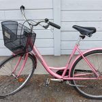 Lohenvärinen, vaaleanpunainen 28-tuumainen naistenpyörä. Pyörässä näkyy useita heijastimia, seisontatuki, etukori, käsijarru ja soittokello.