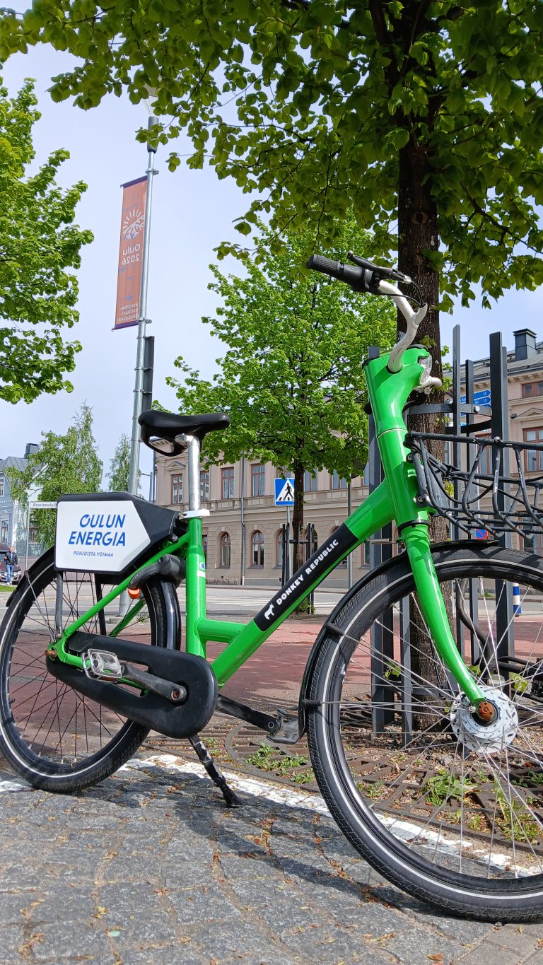 Donkey Republic -merkillä varustettu vihreä kaupunkipyörä parkkeerattuna puun viereen Oulun keskustassa. Pyörässä on näkyvällä paikalla sponsori Oulun Energian logo. Taustalla liehuu tangosta Oulu 2026 viiri.