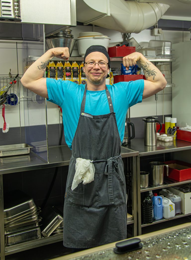 Keittiön työntekijä, joka on kirkkaan vaaleansinisessä t-paidassa ja tummanharmaassa essu, poseraa kameralle kädet ylös sivuille koukkuun nostettuina. Takana taustalla näkyy teräksisinen taso ja hyllyjä, jotka sisältäävät erilaisia keittiötarvikkeita, kuten vuokia, kattiloita, muovikuppeja, mehupurkkeja ja tiskausvälineitä.