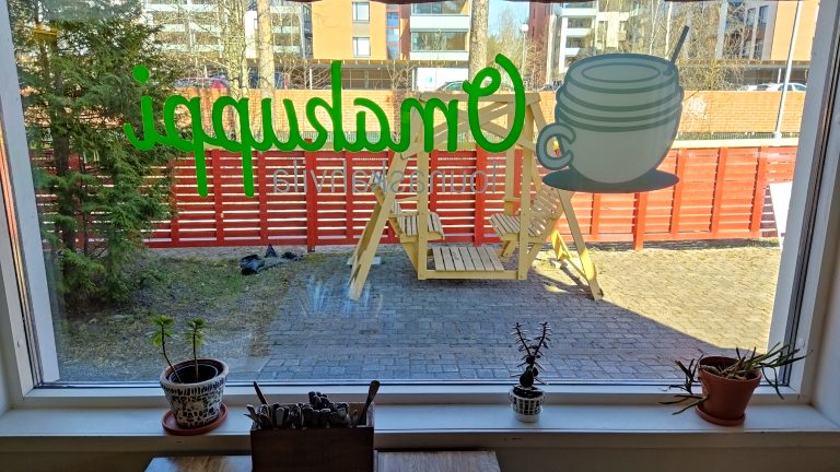 Ikkunalaudalla on kolme pientä huonekasvia ruukuissaan, ja lisäksi ruokailuväline teline on nähtävissä. Ikkunasta katsottuna ulkopuolella näkyy vihreä puu, punainen aita ja keltainen pihakeinu. Aidan takana näkyy oranssi tiiliaita, kerrostaloja ja puiden runkoja.