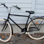 Musta 28 -tuumainen Yosemite Single Speed-merkkinen korkearunkoinen kaupunkipyörä. Pyörässä on useita heijastimia, käsintoimiva etujarrut, seisontatuki, tarakka, krominen soittokello, valkoiset renkaat.