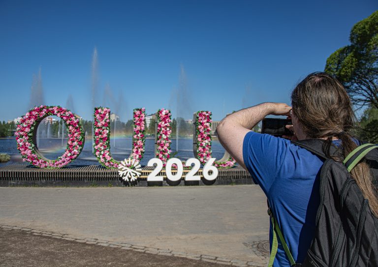 Lava, jossa punaisin, vaaleanpunaisin ja valkoisin kukkasin muotoillut O, U, L ja O -kirjaimet. Alla valkoisella logo ja teksti 2026. Edessä siniseen paitaan pukeutunut tummahiuksinen kuvaa ottava henkilö. Kirjainten takana sininen taivas ja suihkulähteitä.