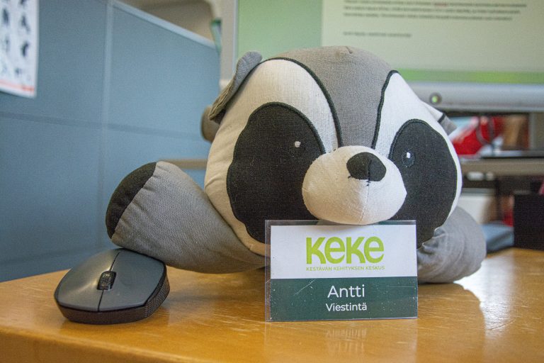 Pesukarhupehmolelu pöydällä. Toisen etujalan alla on tietokoneen hiiri ja leuan alla nimikyltti tekstillä: KeKe, Antti, Viestintä.