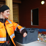 Työllisyyspalvelupäällikkö Rosalie Leskelä seisoo Sorvi-toimipaikan paperinkeräyspisteen vieressä. Rosalla on päällään Kestävän kehityksen logolla varustettu kirkkaan oranssi työtakki.