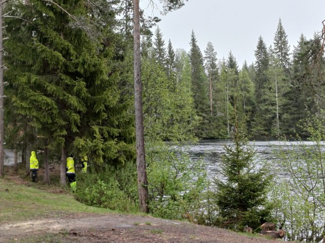Kolme kirkkaan huomiovärisiin työvaatteisiin pukeutunutta henkilöä nojailemassa havupuihin ja katselemassa yhdessä joelle.