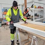 Joni_onnistumistarinaan Viherrakentaja Joni seisoo työpöydän äärellä Hieta-toimipaikan työpajalla. Jonilla on kirkkaankeltaiset huomiovaatteet päällään. Joni mittaa työpöydällä makaavaa ovenkarmea mittanauhalla.