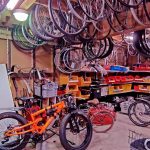 Tarvikevarasto Pyöräkorjaamon tarvikevarasto. Seinät ovat täynnä hyllyjä ja telineitä erinäisille varaosille. Katosta roikkuu renkaita ja vanteita. Lattialla on kunnostettavana olevia polkupyöriä.