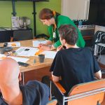 Green Care -ideapaja Kestävän kehityksen keskuksen luokkatilassa. Pöydällä on Green Careen liittyviä papereita, joihin on kiinnitetty useita muistilappuja. Pöydän ääressä istuu kaksi mustapaitaista henkilöä, ja pöydän vieressä seisoo vihreäpaitainen nainen, joka kirjoittaa muistilapulle jotain.